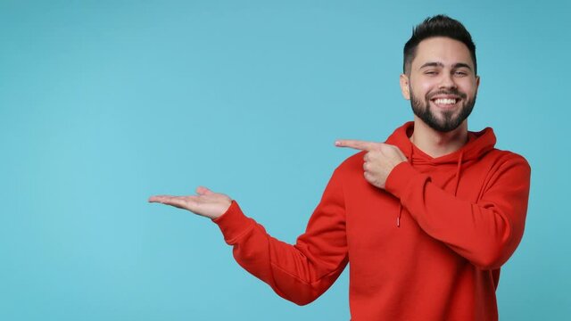 Cheerful funny young man 20s years old in casual red streetwear hoodie isolated on blue background studio. People lifestyle concept. Pointing index finger on empty palm showing thumb up like gesture
