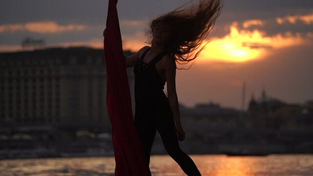 Silhouette of Female dancer showing her flexibility and splits with red aerial silk on the cityscape background during dramatic sky in slow motion. Concept of passion, desire and attraction 