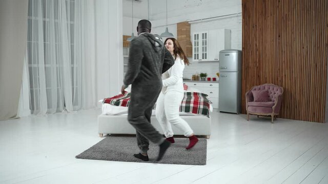 Attractive multinational couple in love wearing fleece pajama suits performing popular slow dance movements during home leisure. Bearded dark-skinned man and his curvy girlfriend dancing together