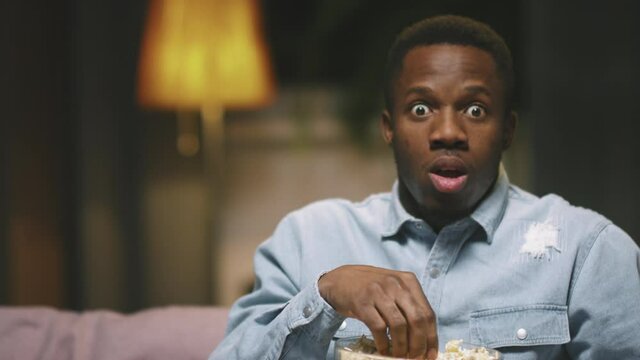 Slow-motion zoom-in waist-up POV of scared young african american man watching horror movie alone at night, eating popcorn while sitting on couch in living room