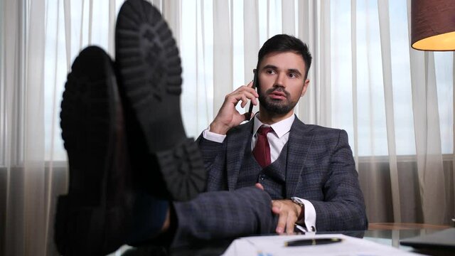 Close-up of respectable businessman sitting with his feet on desk and communicating on cellphone with his wife and telling about business trip delay. Bearded male chatting on smartphone in hotel room