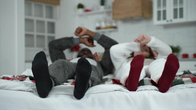 Closeup of feet in socks making dance moves, blurred on background cheerful dark-skinned man and curvy woman wearing kigurumi pajamas performing funny hand and leg movements while lying on bed at home