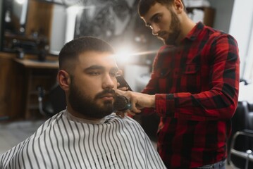 Making haircut look perfect. Young bearded man getting haircut by hairdresser while sitting in chair at barbershop