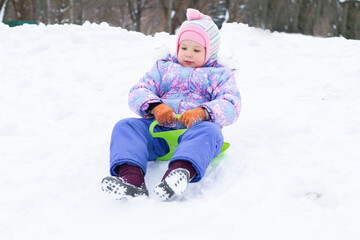 A little pretty girl in a purple overalls slides down an ice slide, winter children's games, a...