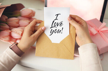 Woman's hands holding a letter in craft envelope. Pink background, valentine's day concept. Tulips flower and pink gift box in background. Womens home desk