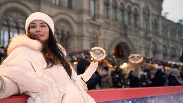 A pretty brunette European girl stands at the side of the ice rink takes pictures looking at the camera and fooling around on the background Christmas market smiling portrait High quality 4k footage