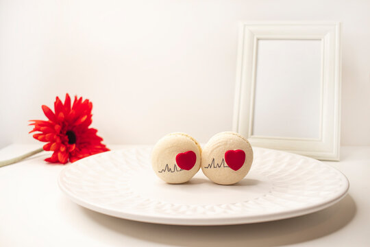 Two gift macaroons with hearts on a white plate, against the background of a photo frame and a red flower. Birthday present, Valentine's Day.