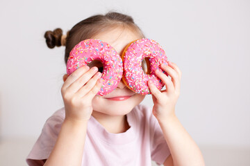 A little girl with blond hair hid behind pink doughnuts and looks through the doughnuts. The joy of the game of hide-and-seek