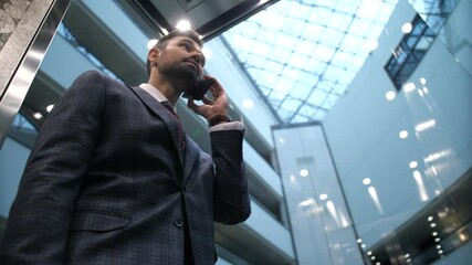 Low angle view of smiling bearded businessman talking on smartphone while going down in transparent lift of luxury hotel. Handsome enterpreneur in business suit during conversation in hotel elevator