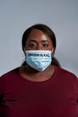 Serious plus size young african american woman in casual clothes wearing protective face mask with Racism is evil text looking at camera, posing isolated over gray background