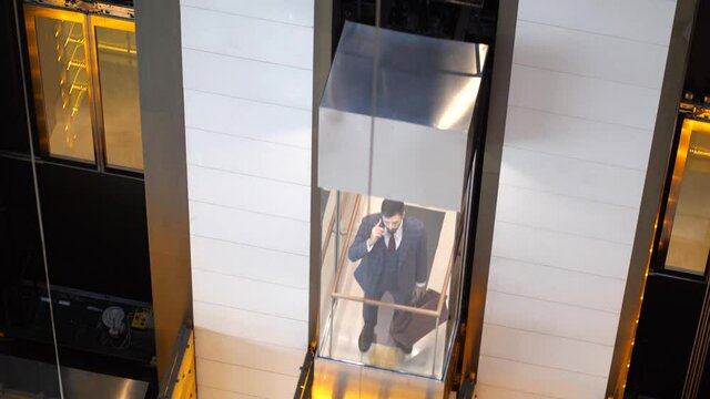 Confident bearded businessman with travel bag talking on cellphone while going up in transparent lift cabin. Respectable hotel guest in chic business suit and tie taking elevator car up to his room