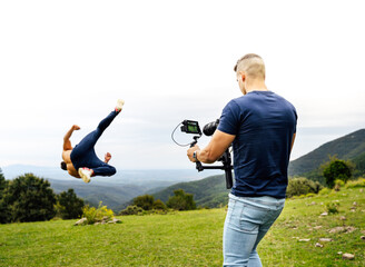 Young and fit cameraman recording with a gimbal a person doing sports, specifically stunts (tricking). Shot from behind, behind the cameras / backstage.