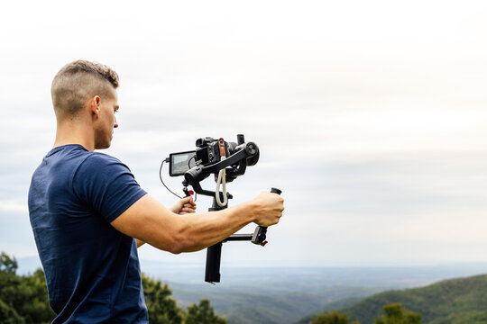 Young muscular Caucasian man manning a camera on top of a gimbal while working recording a scene in the middle of a field. Videomaker with steadicam and professional team working.