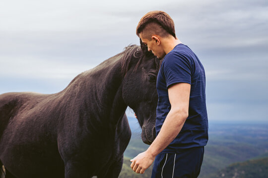 Young Caucasian man with mohawk hair hugging a black horse in the middle of the mountain on a day with cloudy sky.