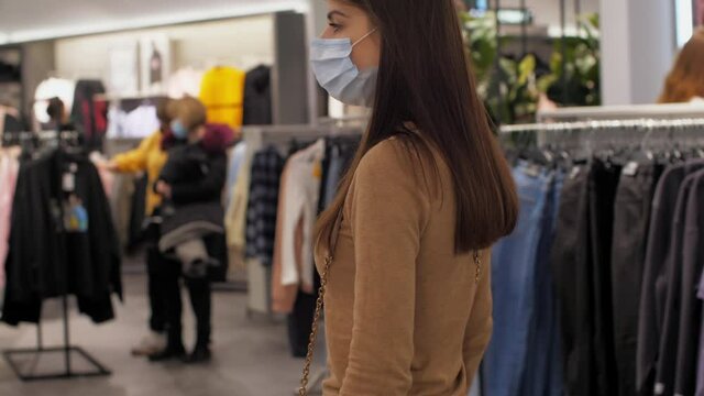 Long haired lady in face mask walks along clothes shop and looks at different pullovers and jeans. Concept coronavirus quarantine