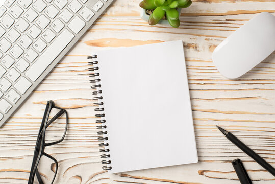 Above overhead close up view photo image of computer mouse flowerpot pen eyeglasses copybook with clean page for mock up on wooden white backdrop