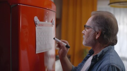 The old man is happy, when he marks the diet schedule hanging on the refrigerator. Old man marking the health calendar hanging on the refrigerator. Diet calendar, chart marking concept