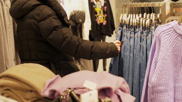 Young woman in disposable mask holds blue jeens in hands and touches materials standing in store