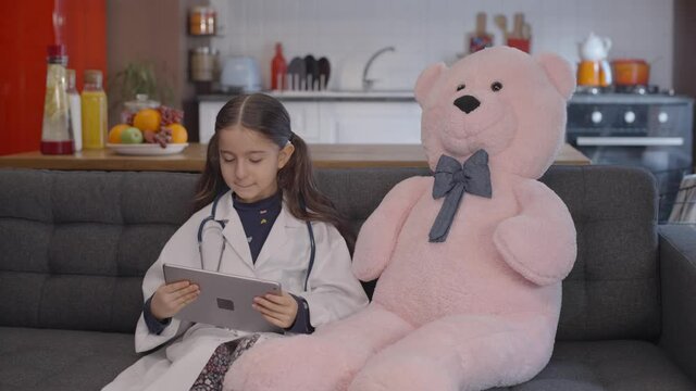 The little girl who tries to play with the pad sits down like a friend with her teddy bear and is happy when she takes scissors from it&rsquo;s cheeck. Teddy bear friend portrait. Game concept with pad.