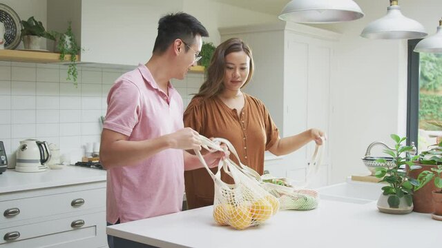 Asian couple unpacking fresh and healthy local fruit and vegetables in zero waste packaging from bag in kitchen at home - shot in slow motion