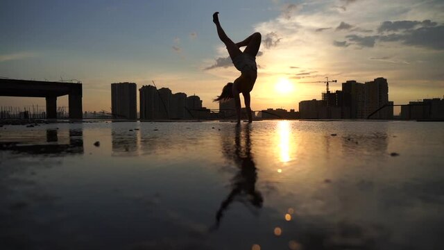 Flexible female gymnast doing handstand and calisthenic with reflection in the water on cityscape background during dramatic sunset in slow motion 