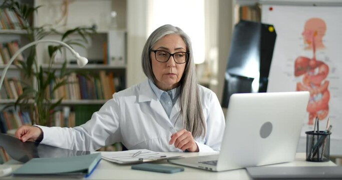 Female doctor holding x ray photo of chest while having online medical consultation in office. Elderly woman in glasses and white rob using laptop while communicating with patient