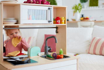 cute toddler baby girl playing on toy kitchen at home, pretends eating slice of pizza