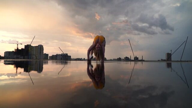 Blond Flexible female gymnast going upside down in the bridge with reflection in the water on cityscape background during dramatic sunset in slow motion 