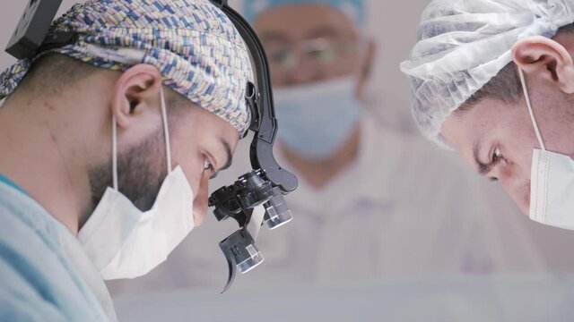 Doctor and an assistant in the operating room for surgical venous vascular surgery clinic. Action. Side view of a surgeon with a binocular loupe.