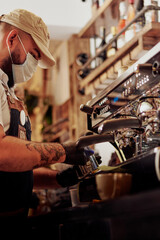 Hombre joven con tatuaje y tapaboca en maquina de expresso sirviendo un cafe