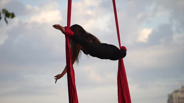 Female acrobat showing her flexibility and splits with red aerial silk on the sky background is slow motion. Concept of grace, movement and beauty 