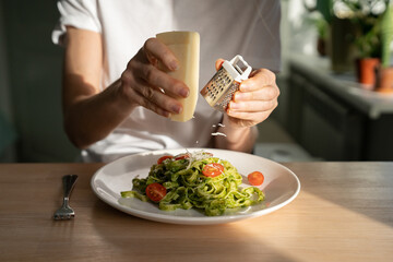 Close up of woman hands grating Parmesan cheese in pasta with sauce pesto, fresh cherry tomatoes, living room on background. Homemade Italian cuisine.