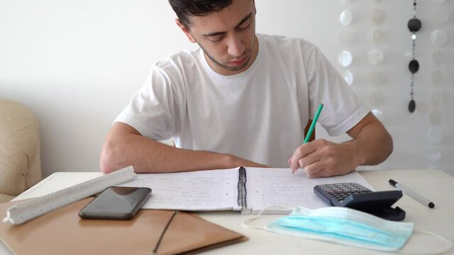 Male Student Writing Mathematical Formulas And Solutions On A Notebook With Calculator, Smartphone, And Medical Mask On The Table. medium shot