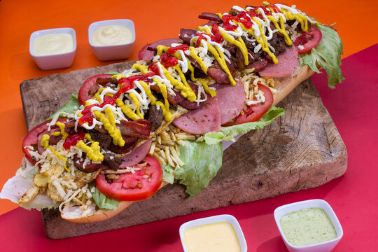 Comida r&aacute;pida venezolana hecho de carne, pollo, lechuga, ensalada de col, tomate, papas al hilo, queso rallado, tomate, tocino con pan baguette, en mesa de madera para restaurante