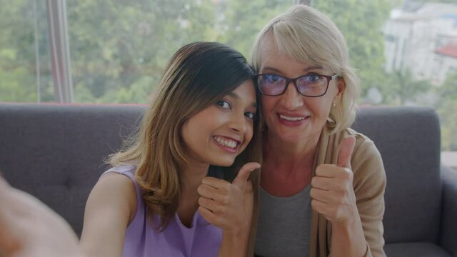 Medium shot - Adopted Asian teen and her Caucasian stepmother enjoy talking a selfie together. Free-time and love sharing of biracial family. 