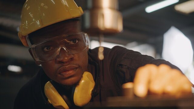 American Industrial Black Young Worker Man With Yellow Helmet And Ear Protection Manual Rotating On Drill Machine In Order To Penetrate Workpiece, Engineer At Work In Industry Factory.