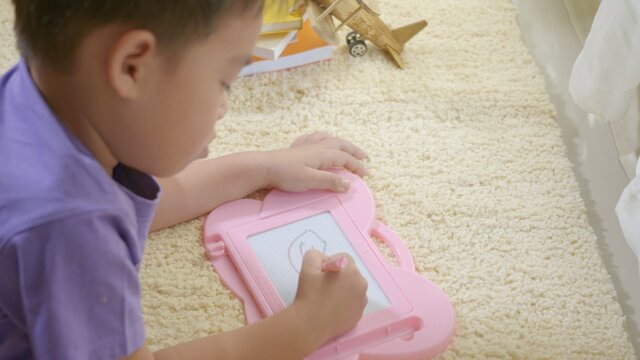 Happy Asian klid little boy preschool writing at the magnetic drawing board at home on carpet. Funny child playing magnetic drawing board. Education Learning to draw concept. Back view