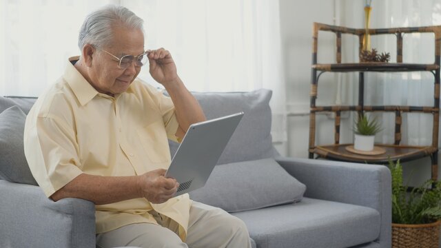 Senior man dressed to wear eyeglasses sitting on sofa looking news on digital tablet computer in living room at home, Happy old man retired using digital technology, Elderly grandfather work from home