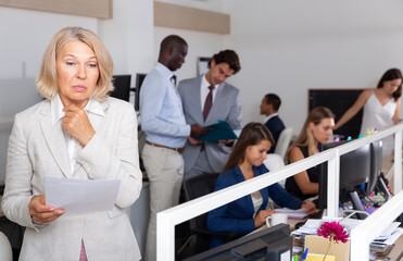 Confused mature female office worker holding papers in office among working colleagues