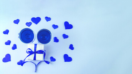 Top view of two glasses with blue wine, gift with blue ribbon and blue hearts on light blue background, banner, copy space. Happy Valentine's Day greetings.