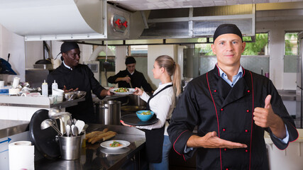 Confident chef of restaurant posing in kitchen on background with working employees..