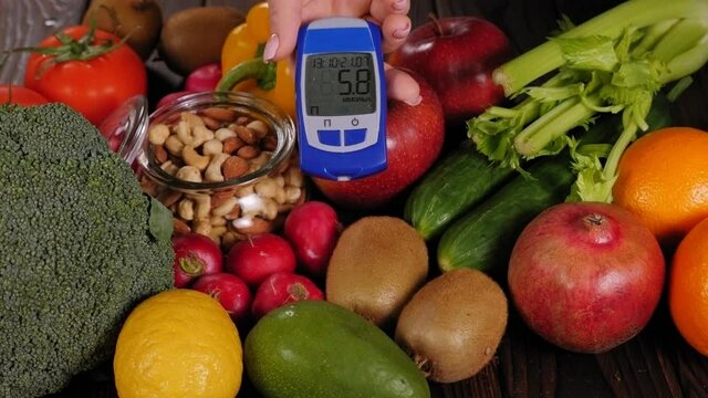 Close-up of many different fruits and vegetables on a wooden table, a woman's hand holding a blood sugar meter to measure blood sugar with a good result.
