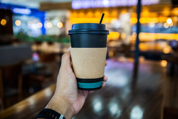 hand holding black carton coffee cup with sleeve paper cup on wooden table with blurred indoor coffee shop background