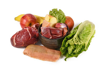 Dog bowl filled with biologically appropriate raw food containing meat chunks, fish, fruits and vegetables surrounded by ingredients on white background