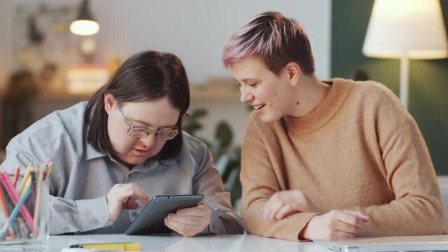 Female social worker sitting with adult man with Down syndrome at table in living room and explaining him how to use digital tablet