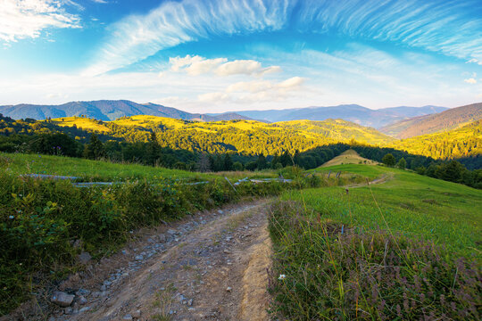 rural landscape in mountains at summer sunrise. country road through grassy pasture winding down in to the distant valley. clouds on the blue sky above the ridge in the distance