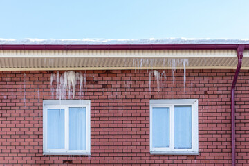 Transparent icicles hang from the snow-covered roof of a brick house. Spring melting snow. Icicles falling danger concept