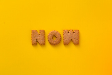 Word Now in the middle of the picture made of tasty crunchy cookies in form of English alphabet letters, textured bright yellow background, health, dieting and medical concept. Copy space, big letters