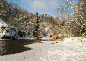 sunny day, landscape with red sandstone cliffs that are snowy with snow, icefall at the cliff wall, frozen river, Gauja, Kuku cliffs, Latvia