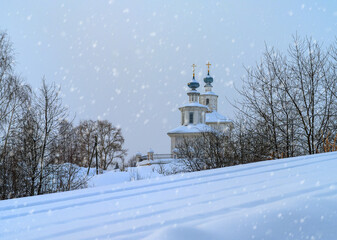 View of a rural church with blue domes on a winter day during a snowfall. In the foreground is a large layer of snow, on which ski tracks are visible. Tall trees are covered with snow 